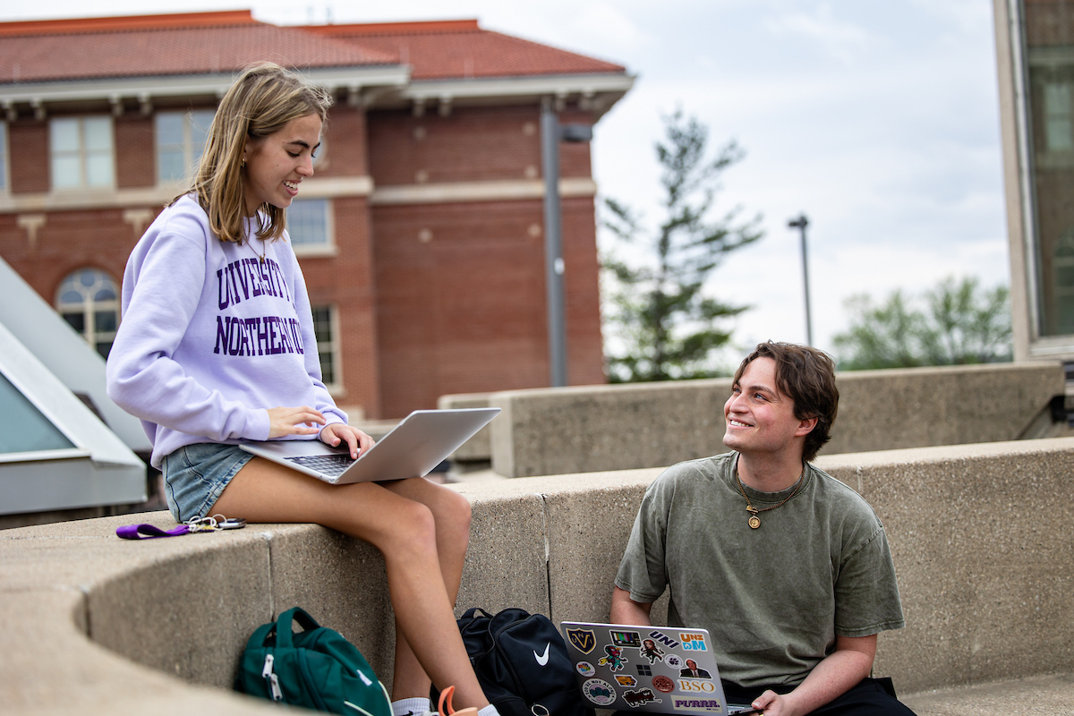 Two college students smile and chat while studying outdoors on a concrete courtyard. A woman sits on a ledge using a laptop, while a man sits on the ground nearby looking up at her with his own laptop open.